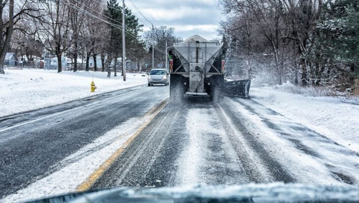 A snowplow truck spreads salt on a snowy road lined with trees during winter