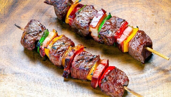 Two skewers with grilled meat and colorful vegetables on a wooden surface