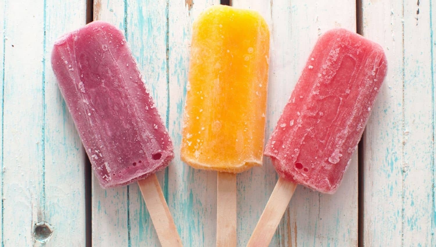 Three colorful ice pops on wooden sticks, placed on a light blue wooden surface