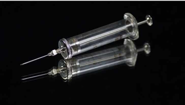 A glass syringe with metal fittings, reflecting on a black surface, invented by Letitia Geer