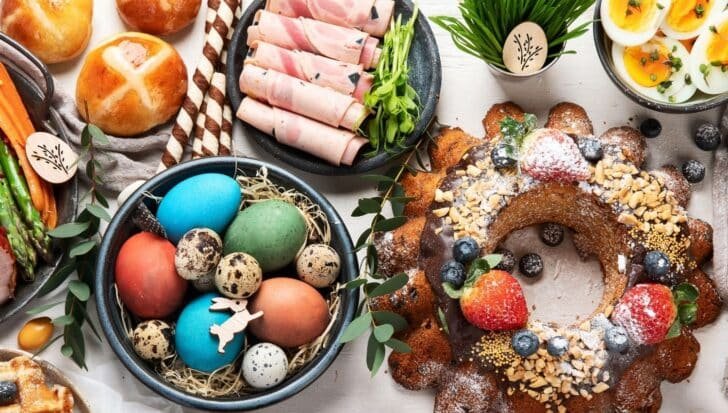 An assortment of Easter foods including hot cross buns, colorful dyed eggs with quail eggs, a ring cake topped with berries and nuts, sliced ham, and a bowl of sliced boiled eggs
