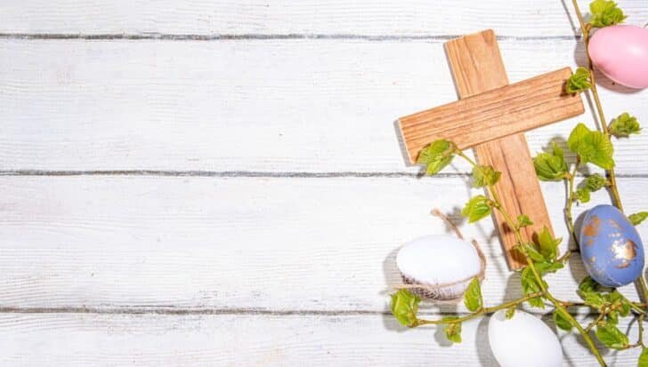 Wooden cross with decorated Easter eggs and green sprigs on a white wooden background