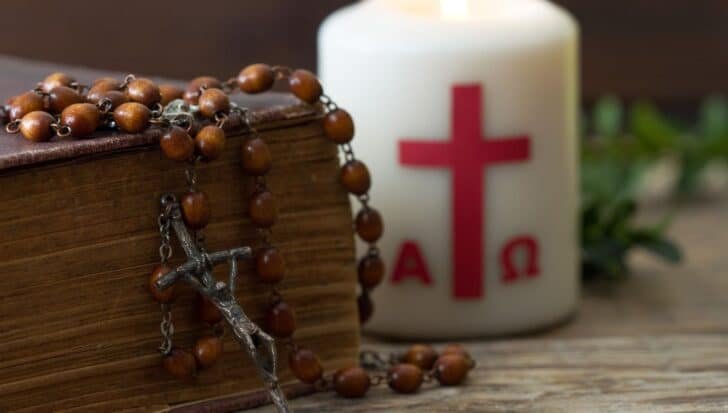 Rosary beads draped over a textured book next to a lit candle with a red cross and Greek letters