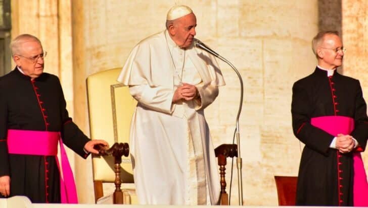 The Pope stands at a microphone dressed in white, flanked by two clergy members wearing black and red robes during Palm Sunday Mass
