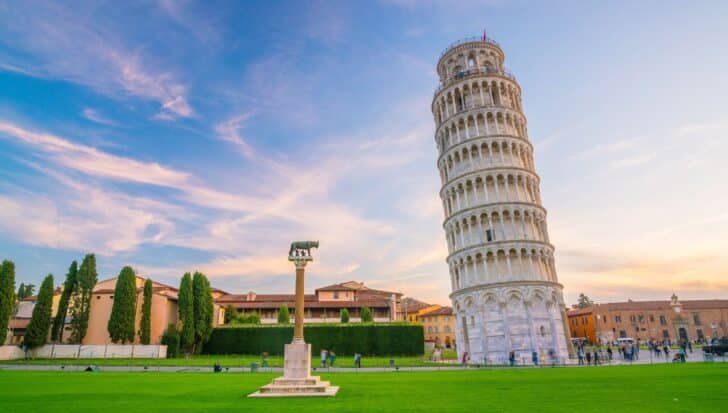 The Leaning Tower of Pisa standing at an angle against a backdrop of a clear sky, surrounded by green grass and nearby buildings