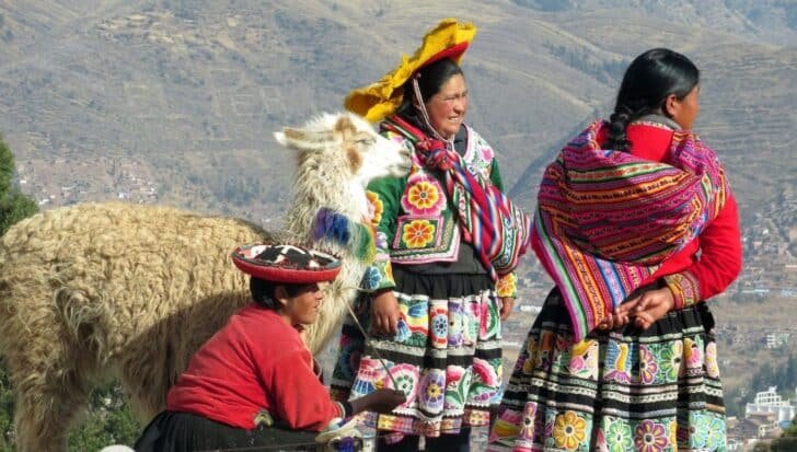 Three women in traditional colorful clothing with a llama, standing in a mountainous region in Peru