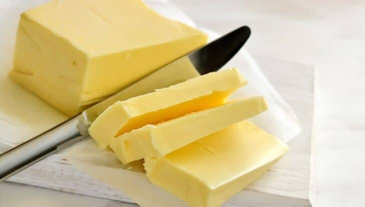 A block of butter with several slices cut from it, placed on a piece of parchment paper with a butter knife