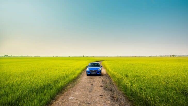 A blue car drives down a dirt road surrounded by vibrant green rice fields under a clear sky.