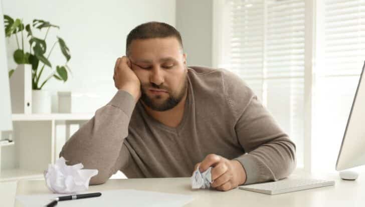 A person looking bored and disengaged at a desk, surrounded by a computer, crumpled paper, and a pen.