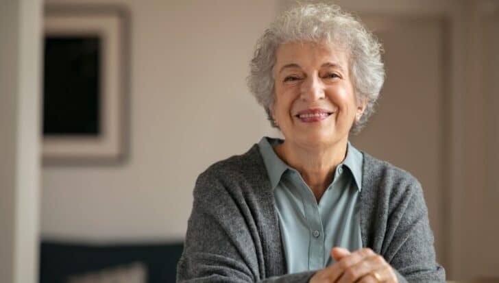 Smiling older woman with curly gray hair wearing a gray sweater over a light blue shirt