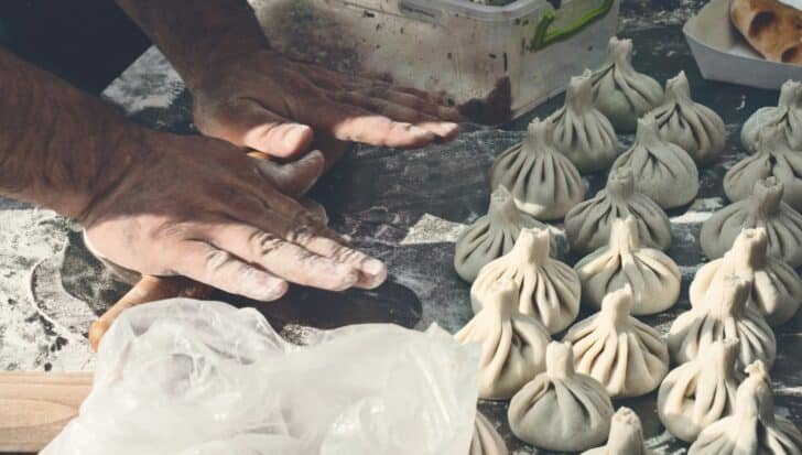 Hands shaping dough on a floured surface with multiple uncooked Georgia Khinkali nearby