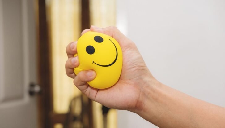 A hand squeezing a yellow stress ball with a smiley face