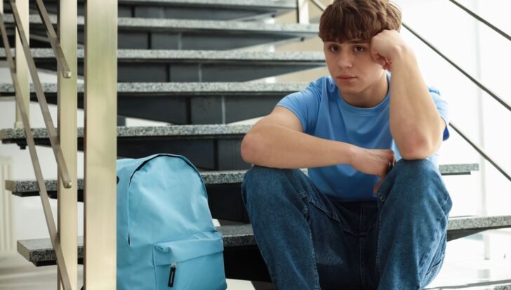 Young person sitting on stairs looking thoughtful with a blue backpack nearby