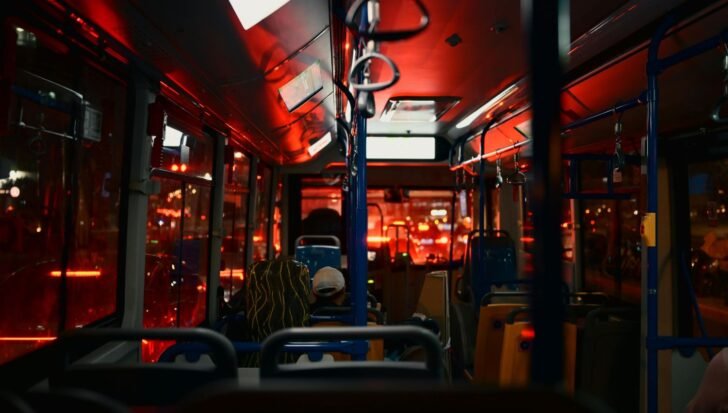 Interior view of a dimly lit bus with several empty seats and a few passengers, red lights reflecting on the windows