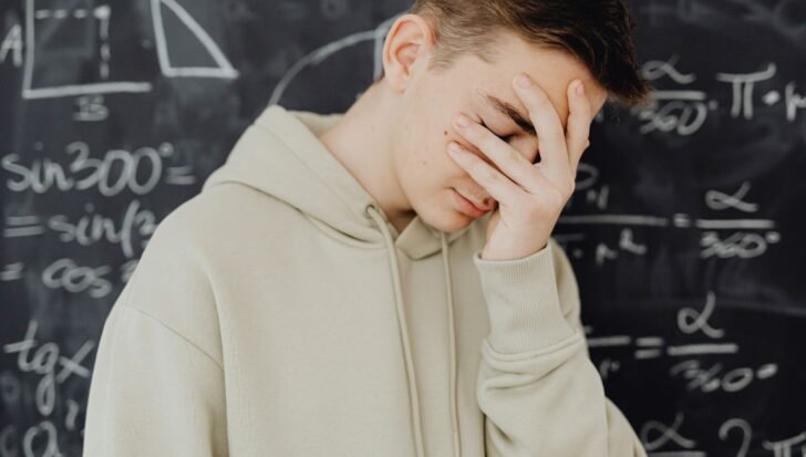 A young man in a hoodie holding their face in frustration, standing in front of a chalkboard