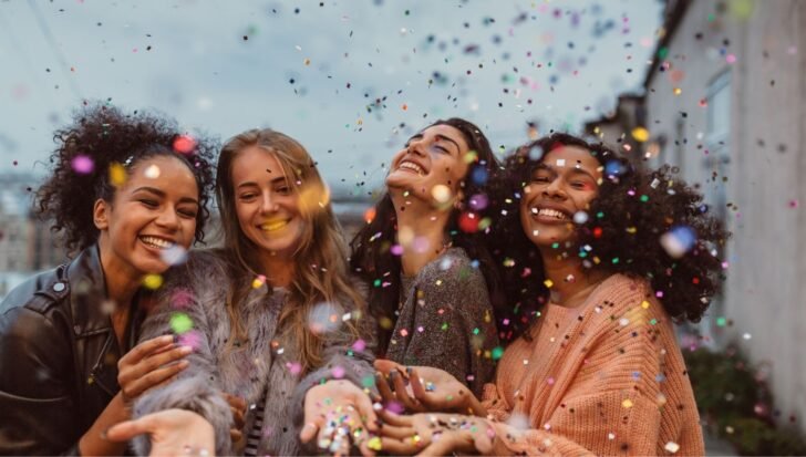 Four women smiling and celebrating under colorful confetti outdoors